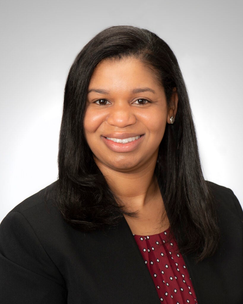 Woman with long, dark brown hair wearing a black blazer over a brown blouse with black and white polks dots.