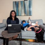 A woman with black hair wearing a black cardigan over a gray top sits on a couch next to two children who are wearing headphones and holding tablets.