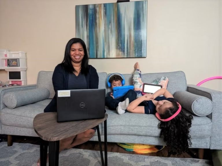 A woman with black hair wearing a black cardigan over a gray top sits on a couch next to two children who are wearing headphones and holding tablets.