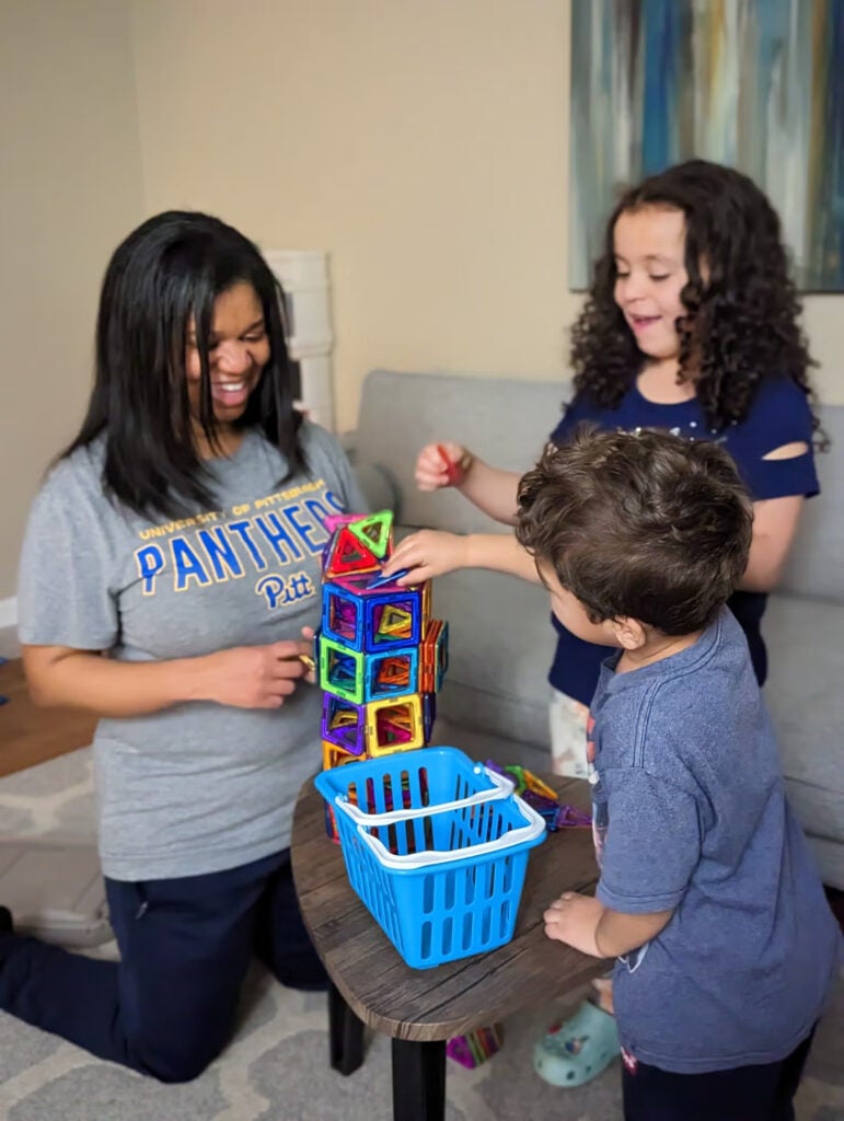 A woman with black hair wearing a gray shirt and jeans kneels on the floor to play with two children -- a young boy and a young girl.