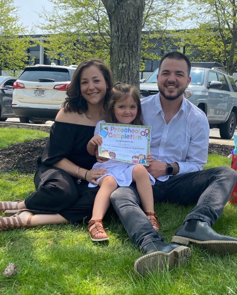 A woman with brown hair wearing a black dress and a man with short dark hair wearing a white collared shirt and black pants sitting in the grass holding a young girl who is wearing a white dress and holding a piece of paper.