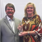 A man with short brown hair and a mustache wearing a tan suit jacket over a white collared shirt standing next to a woman with blonde hair wearing a brown and orange patterned dress.