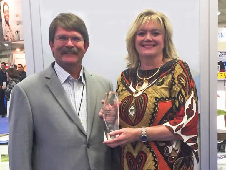 A man with short brown hair and a mustache wearing a tan suit jacket over a white collared shirt standing next to a woman with blonde hair wearing a brown and orange patterned dress.