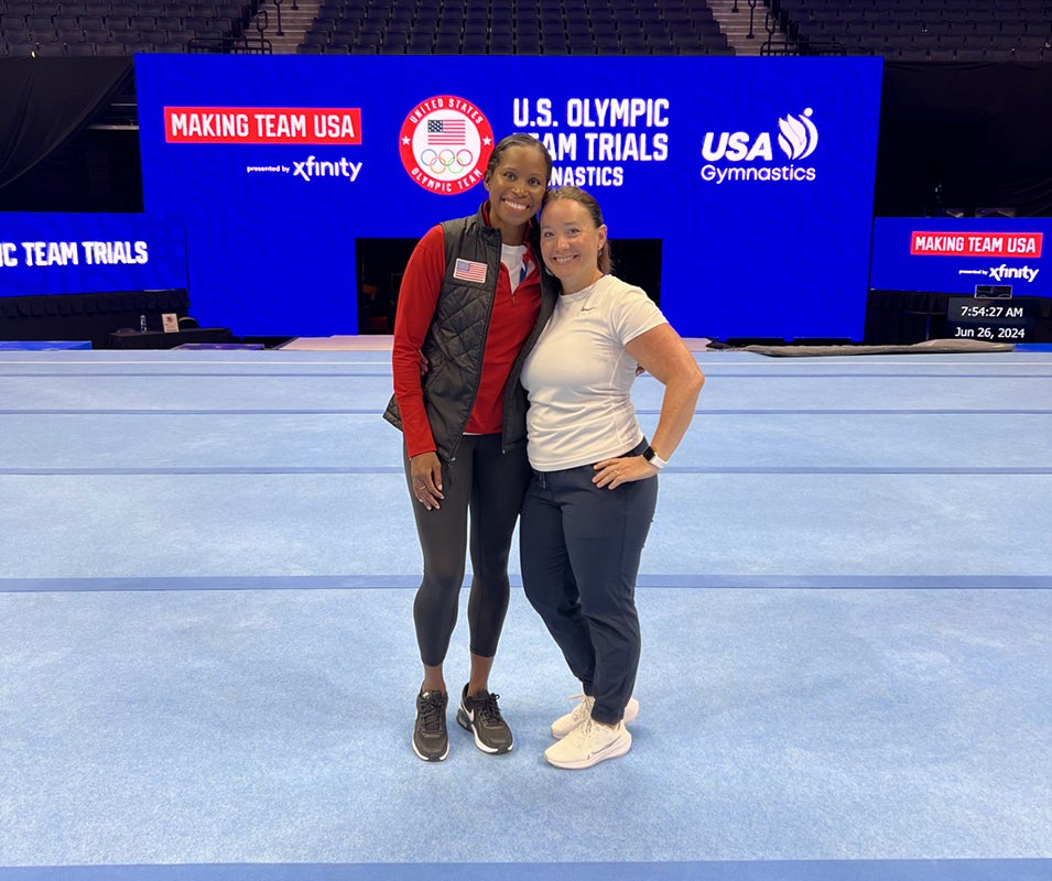 Two women stand together smiling in front of a backdrop for the U.S. Olympic Team Trials and USA Gymnastics