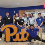 A group of men and women wearing collared shirts and polos standing together in from of a blue and gold "Pitt" sign.