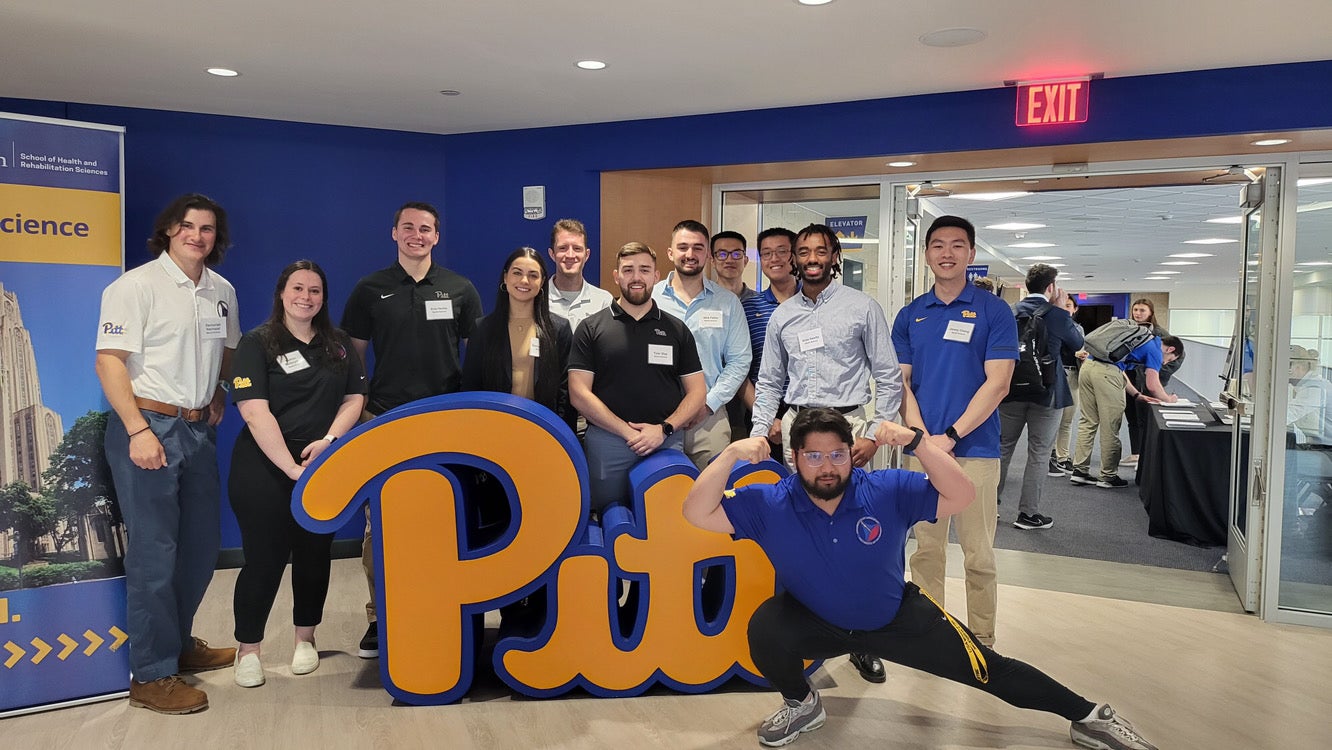 A group of men and women wearing collared shirts and polos standing together in from of a blue and gold "Pitt" sign.