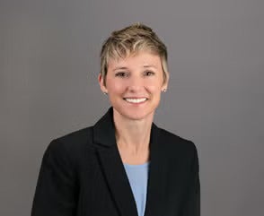 Woman with short brown and blonde hair wearing a black blazer over a light blue top.