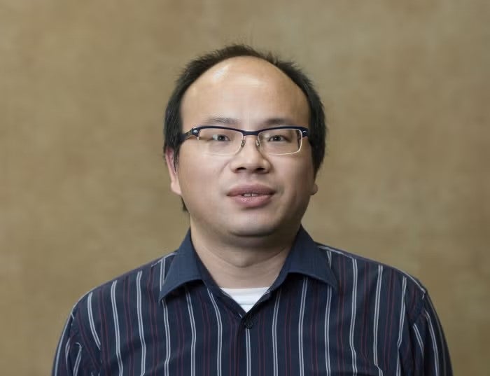 A man with short black hair and black-rimmed glasses wearing a navy blue collared shirt with white stripes.