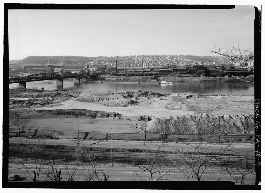 A black and white photo shows an empty dirt parcel in the foreground and a decommissioned steel mill on the other side of the river.