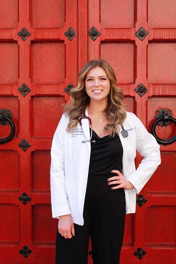 Woman with dark blonde hair wearing a white lab coat over a black jumpsuit with a stethoscope around her neck.