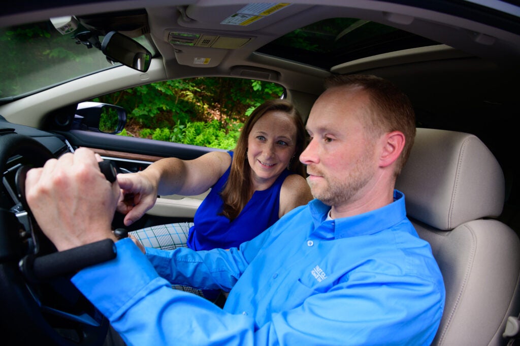 A man and a woman sitting in a car. The man is in the foreground with short light brown hair wearing a long sleeve light blue collared shirt and the woman in the background has brown hair wearing a dark blue tank top and plaid pants pointing to adaptive driving equipment.