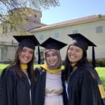 Three women standing together wearing black graduation gowns and caps with gold sashes.