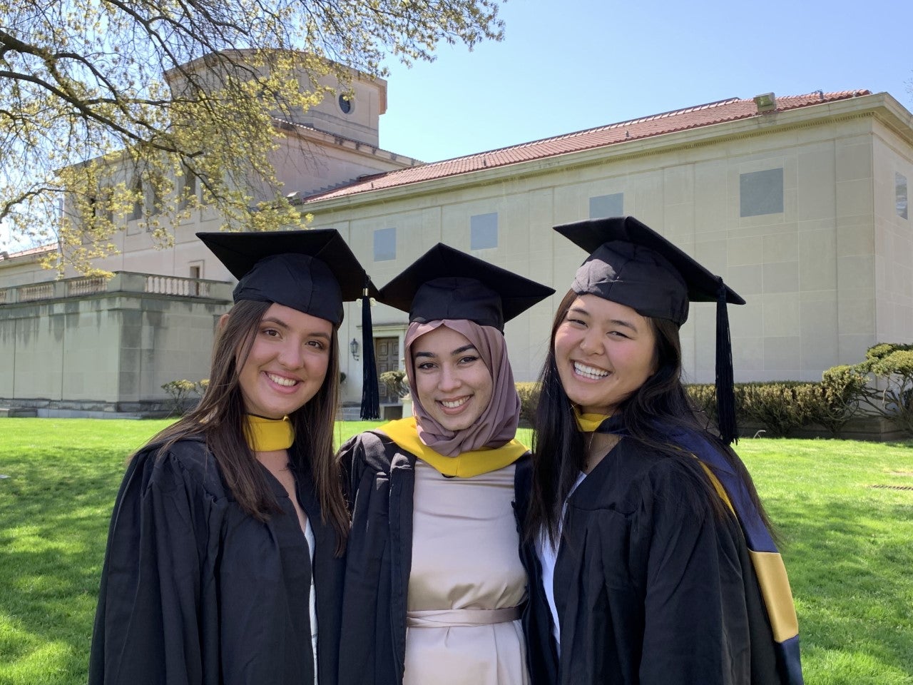 Three women standing together wearing black graduation gowns and caps with gold sashes.