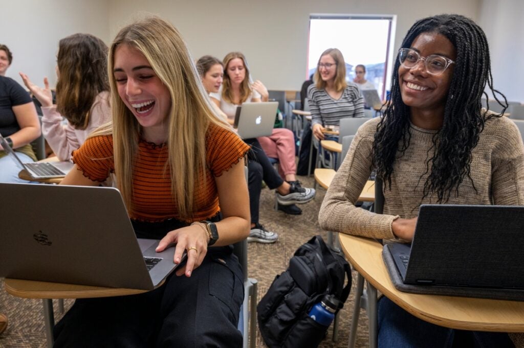 A woman with blonde hair wearing an orange shirt and black pants and a woman with black braided hair wearing a tan sweater and jeans sitting at the front of a classroom together with their laptops, while other students chat in their seats in the back.