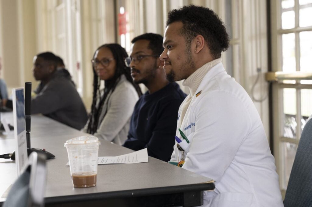 A man with dark brown hair wearing a white lab coat over a tan turtleneck sits at the head of a long table for a panel.