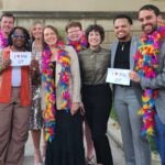 A group of people standing together outside while wearing colorful feather boas and holding signs that read "I heart Pitt OT."