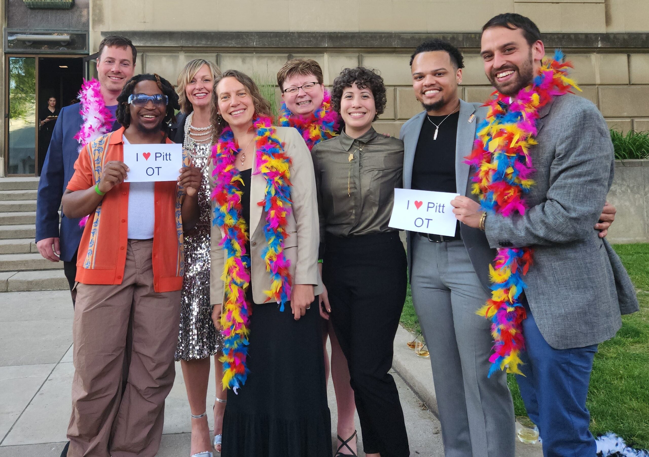 A group of people standing together outside while wearing colorful feather boas and holding signs that read "I heart Pitt OT."