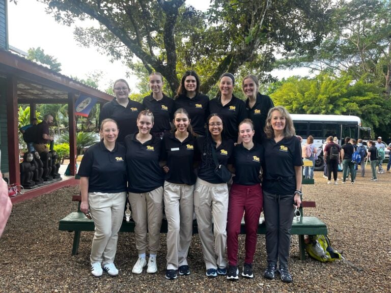 A group of 11 women wearing dark Pitt shirts and light and dark pants gathered together in front of a bench.