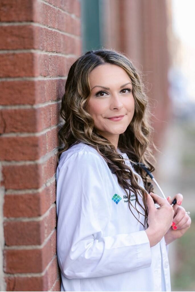 Woman with curled brown hair wearing a white lab coat in front of a brick wall.