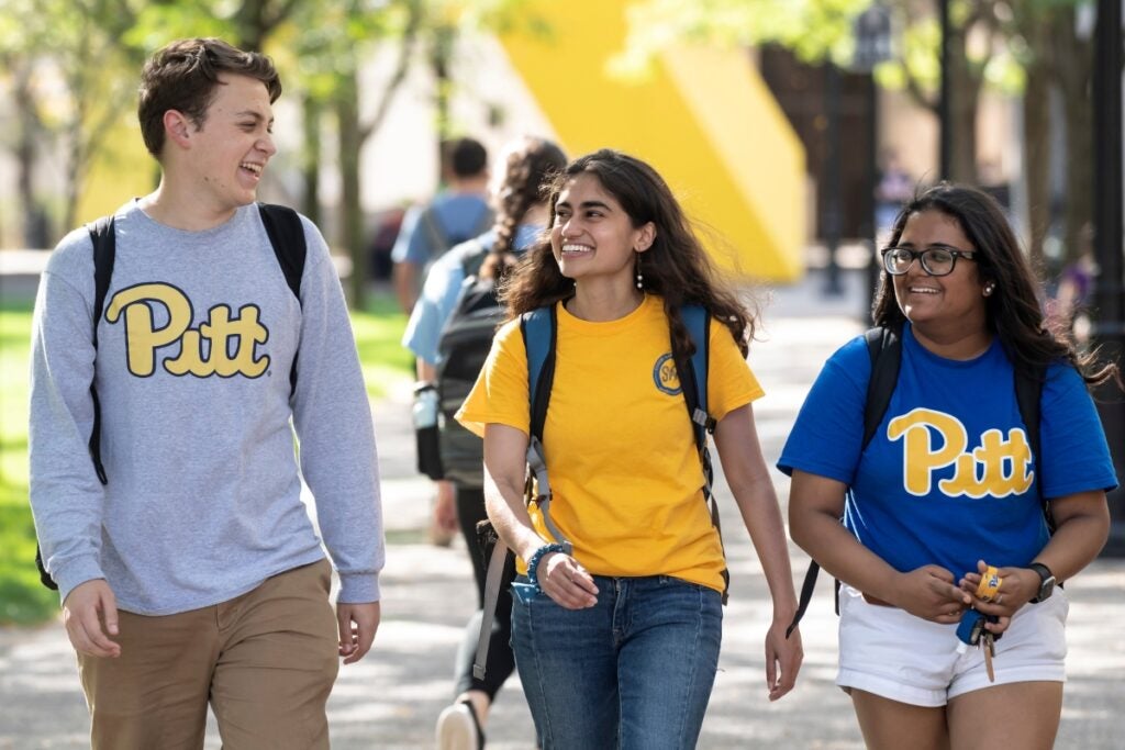 One male and two female students walking together on campus. All three are wearing Pitt gear and backpacks while smiling at each other.