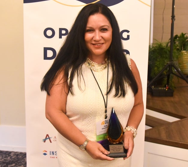 Woman with long dark hair wearing a white dress with white patterned detailing, a pearl necklace and a silver watch while holding a teardrop-shaped award.