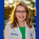 A woman with light red hair wearing glasses and a white lab coat over a green shirt.