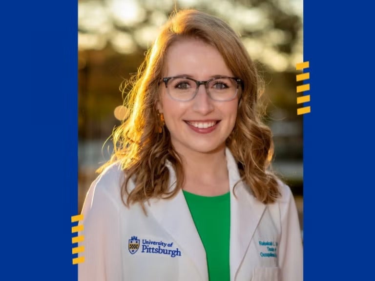 A woman with light red hair wearing glasses and a white lab coat over a green shirt.