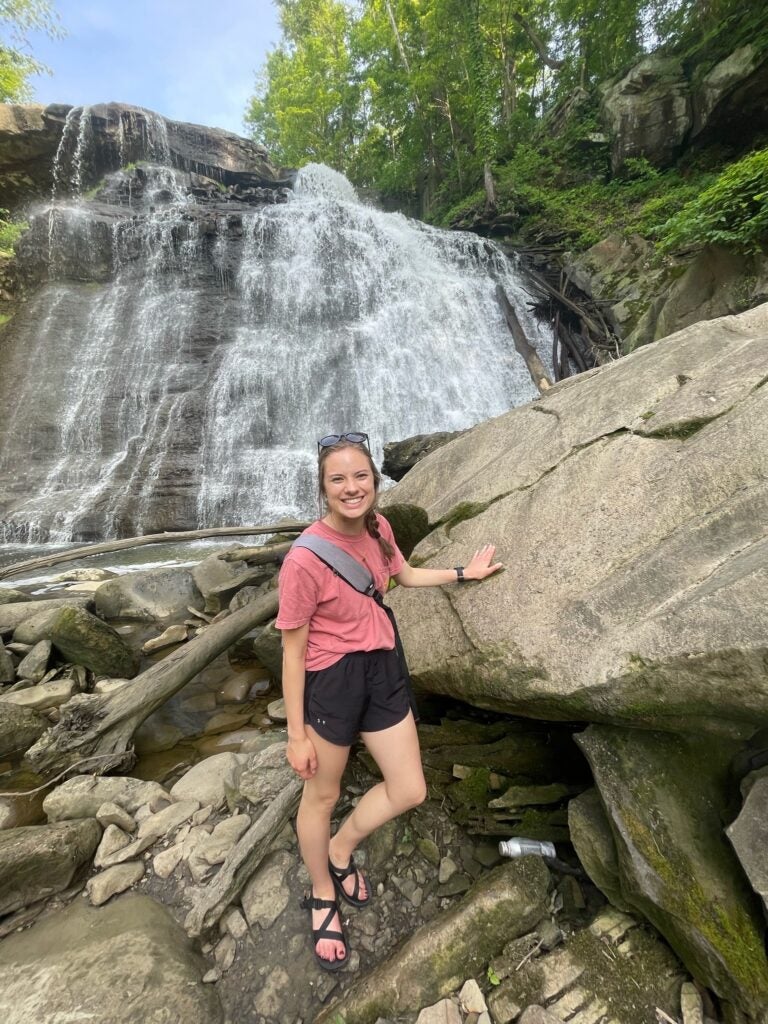 One woman with her hair in a braid wearing a pink shirt and black shorts near a waterfall.