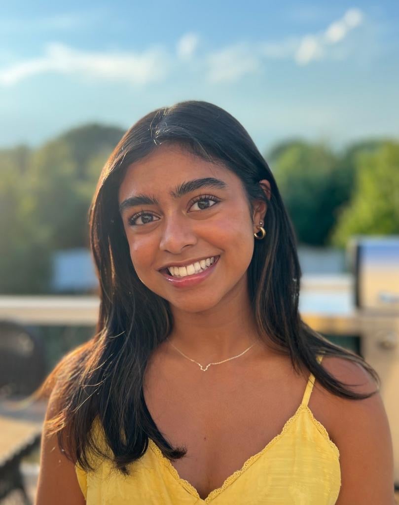 A woman with dark brown hair wearing a yellow top, gold earrings and a gold necklace.