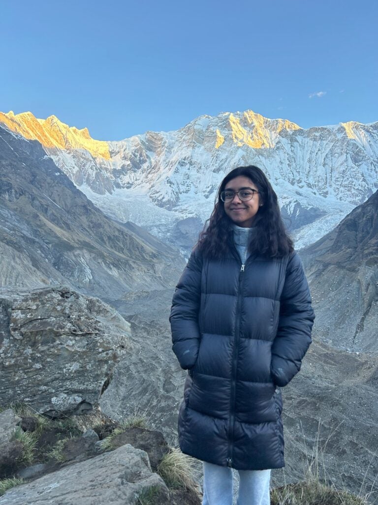 A woman with dark brown hair wearing glasses and a long black puffer coat standing in front of the Himalayan Mountains.