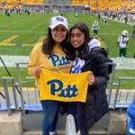 Two brunette women standing next to each other wearing University of Pittsburgh shirts and hats and holding a yellow Pitt towel in front of a football field.