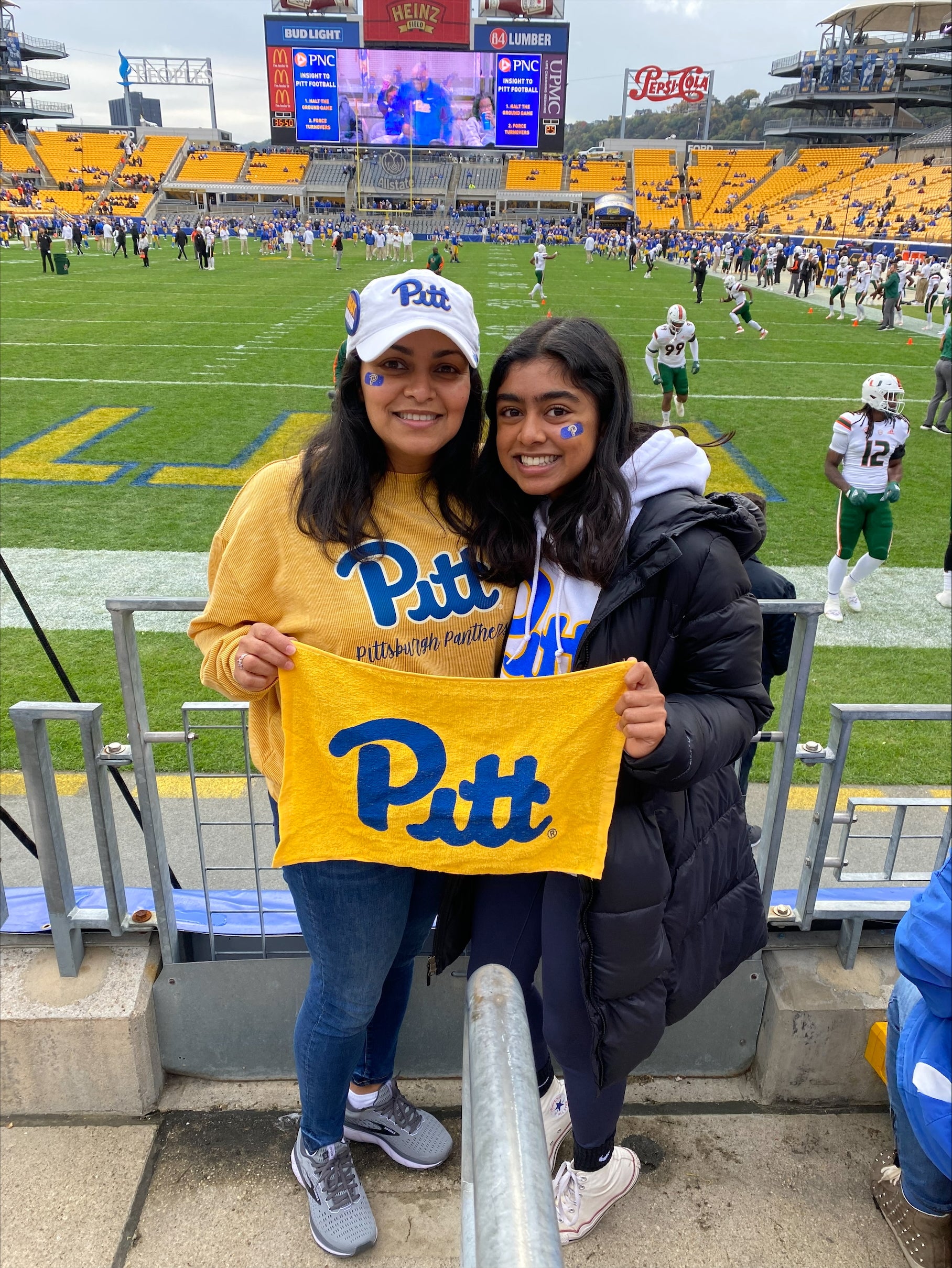 Two brunette women standing next to each other wearing University of Pittsburgh shirts and hats and holding a yellow Pitt towel in front of a football field.