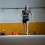 A woman wearing a gray t-shirt and black shorts running in a fieldhouse.