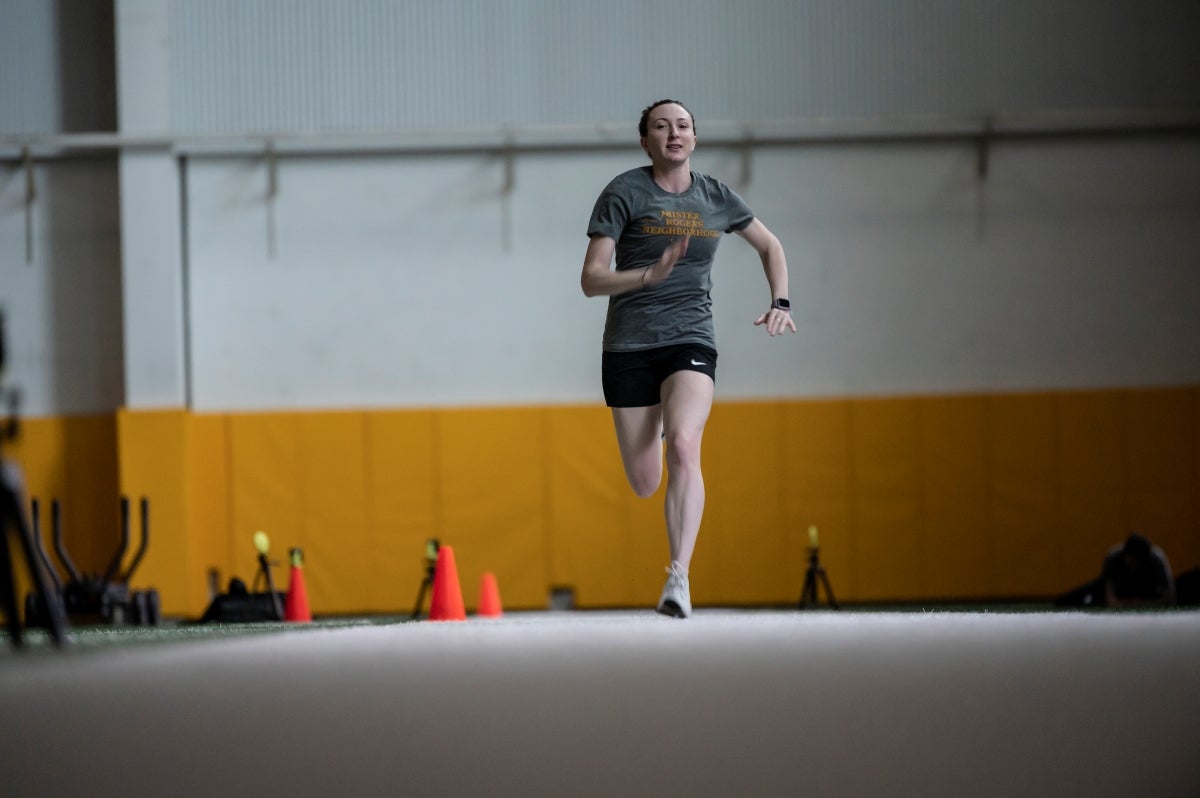 A woman wearing a gray t-shirt and black shorts running in a fieldhouse.