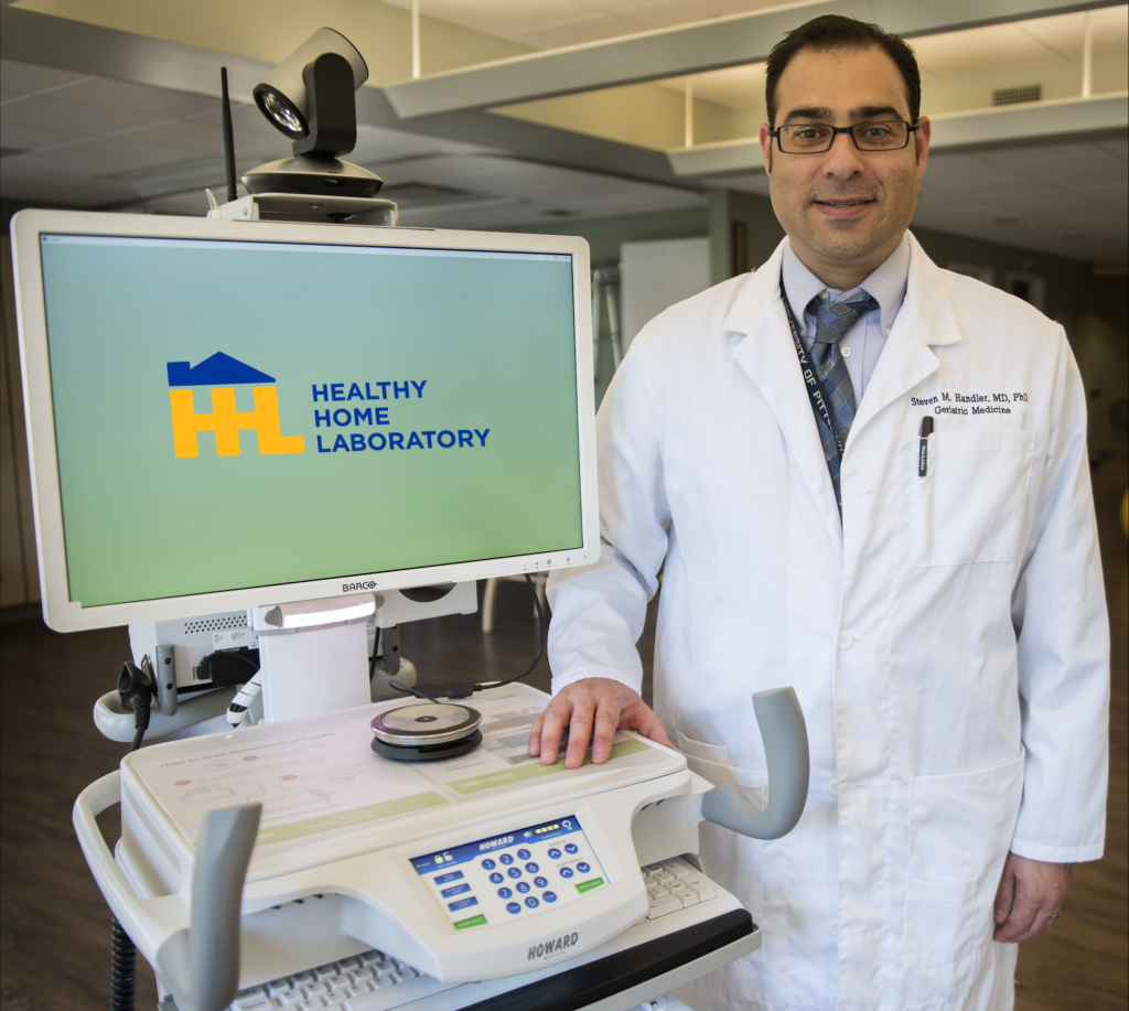 A man with short dark hair and glasses wearing a white lab coat over a light gray collared shirt and gray patterned tie standing next to a screen that reads "Healthy Home Laboratory."