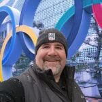 Ed Strapp standing in front of a sculpture of the Olympic rings with a big smile and snowflakes on his hat and jacket