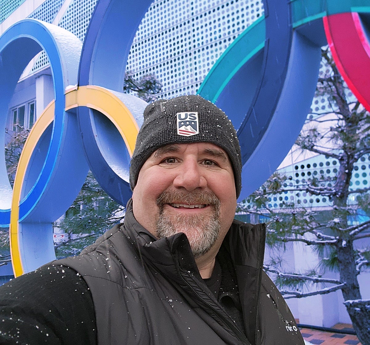 Ed Strapp standing in front of a sculpture of the Olympic rings with a big smile and snowflakes on his hat and jacket