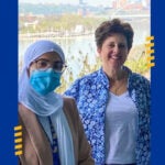 Woman wearing a white hijab, brown glasses, a face mask, a tan blazer, a white top and a Pitt lanyard standing next to a woman with short brown hair wearing a blue floral overshirt in front of the Pittsburgh skyline.