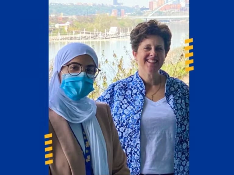 Woman wearing a white hijab, brown glasses, a face mask, a tan blazer, a white top and a Pitt lanyard standing next to a woman with short brown hair wearing a blue floral overshirt in front of the Pittsburgh skyline.