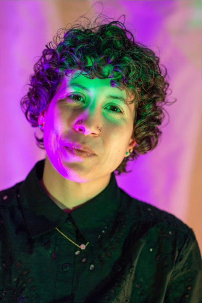 A person with dark, short curly hair wearing a black collared shirt and a gold necklace against colorful lighting that is reflected on their face.