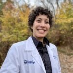 A person with dark, curly short hair wearing a white lab coat over a black collared shirt with gold detailing.