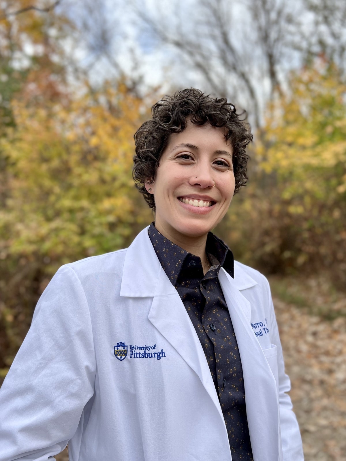 A person with dark, curly short hair wearing a white lab coat over a black collared shirt with gold detailing.