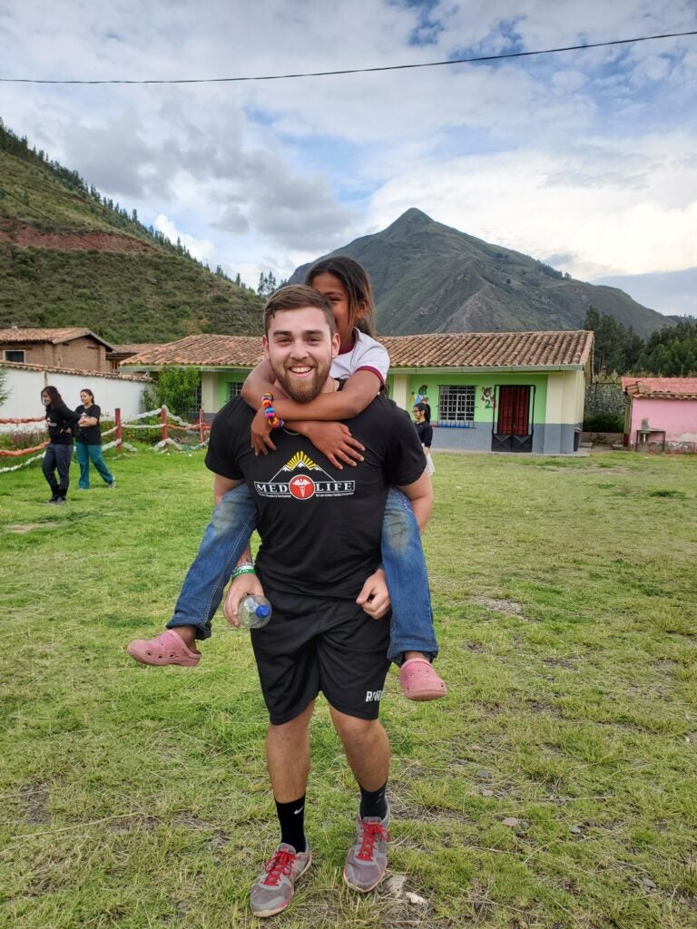 A man with short auburn hair and a beard wearing a black t-shirt and black shorts while carrying a little girl on his back who has brown hair and is wearing a white shirt and jeans.