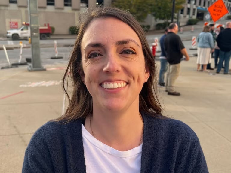 Woman with brown hair wearing a navy blue sweater over a white shirt smiling outside.