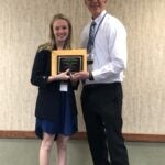 A woman with blonde hair wearing a black cardigan over a dark blue dress standing next to a bald man wearing a white collared shirt and black pants while they both hold a plaque.