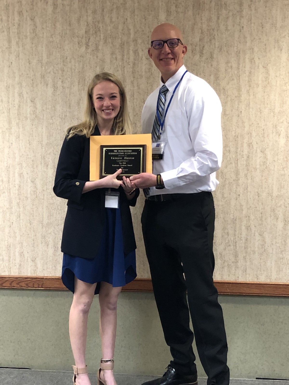 A woman with blonde hair wearing a black cardigan over a dark blue dress standing next to a bald man wearing a white collared shirt and black pants while they both hold a plaque.