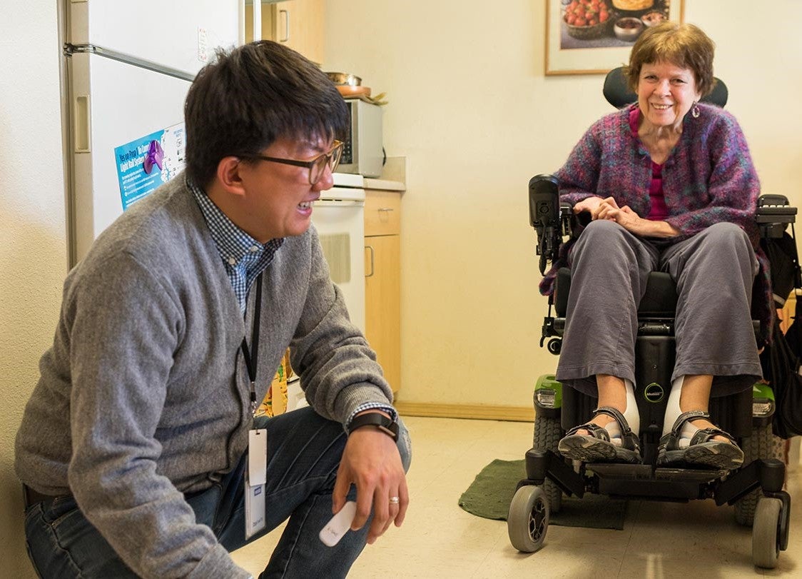 A man with short black hair wearing a gray sweater and jeans kneels down to laugh near a woman who is seated in a motorized chair, wearing a purple patterned sweater and dark gray pants.