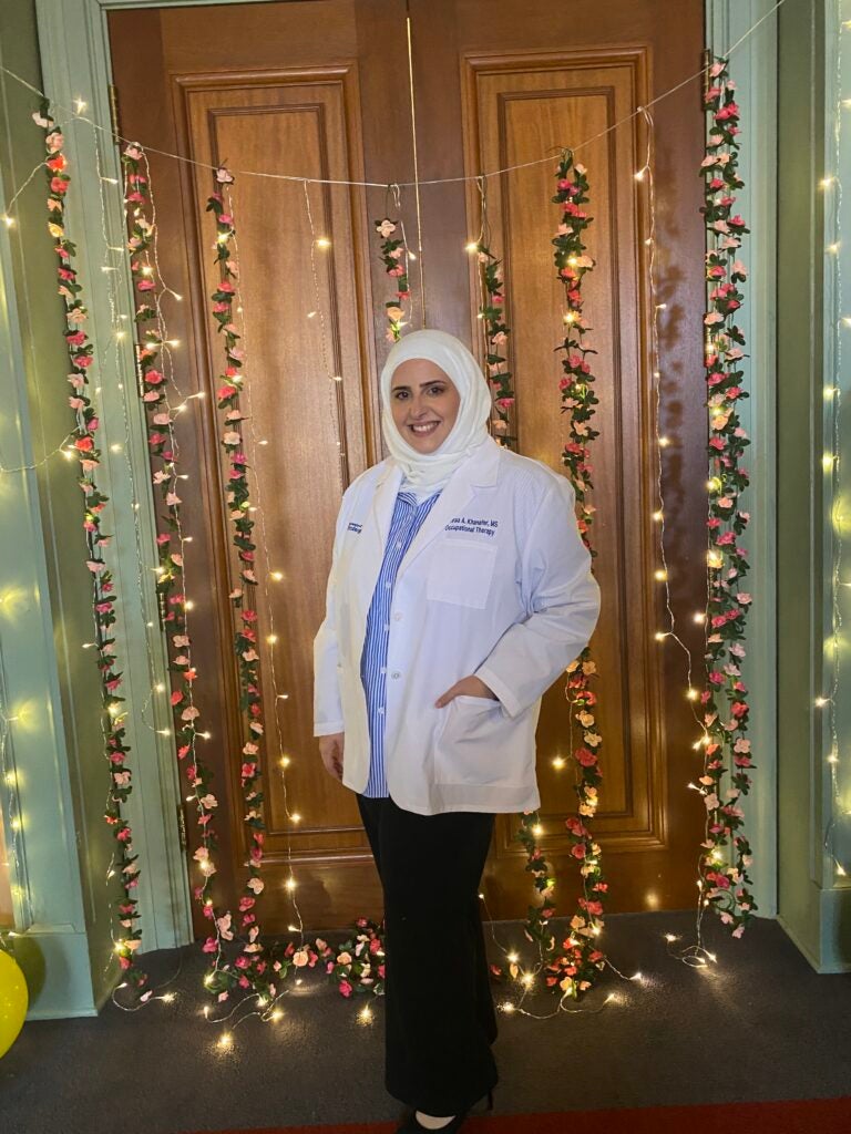 Woman wearing a white hijab, black pants, a blue and white striped blouse top and a white lab coat standing in front of a wooden door with string floral lights.