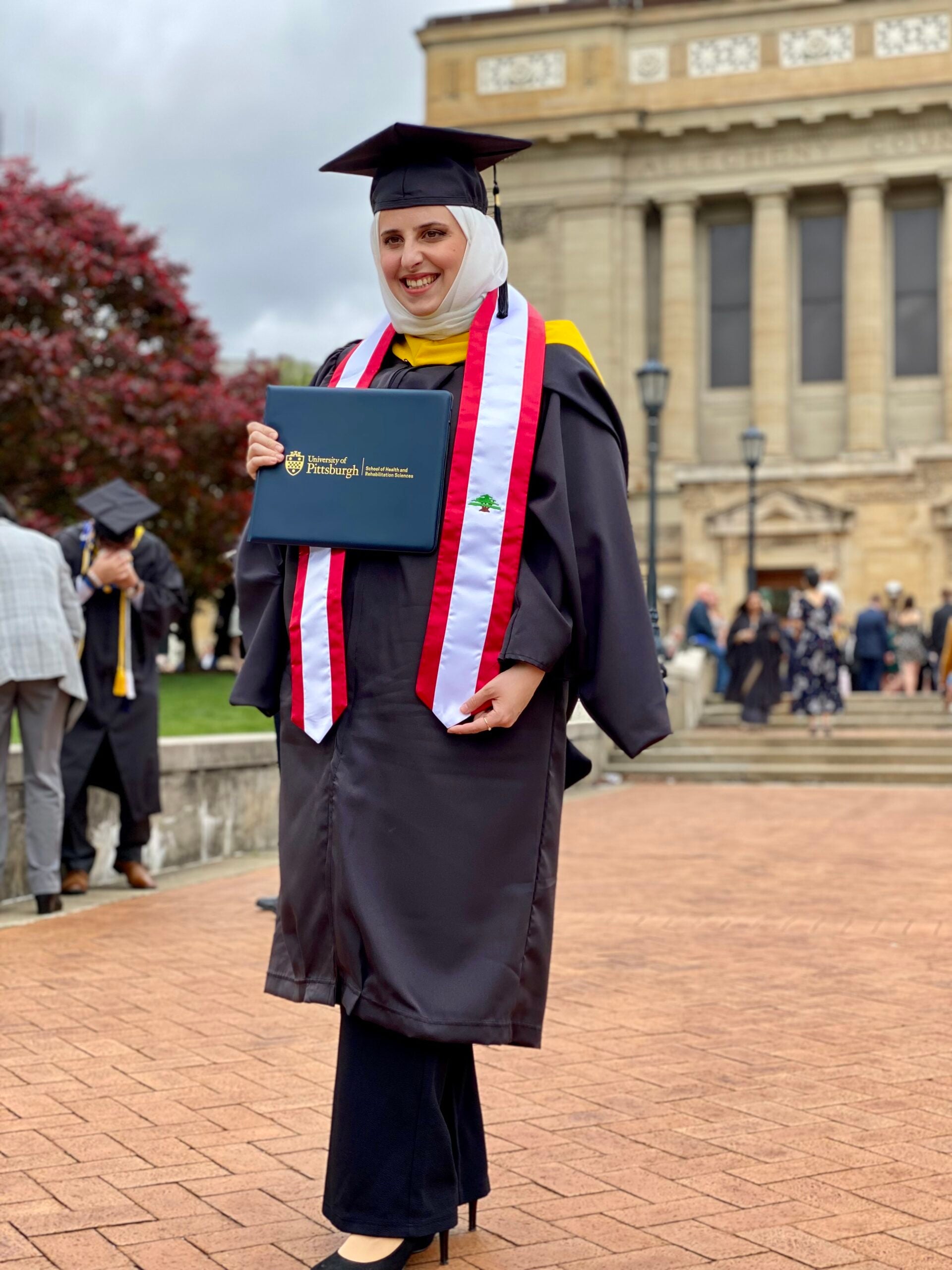 Woman wearing a white hijab and black pants in her graduation cap and gown holding her diploma.